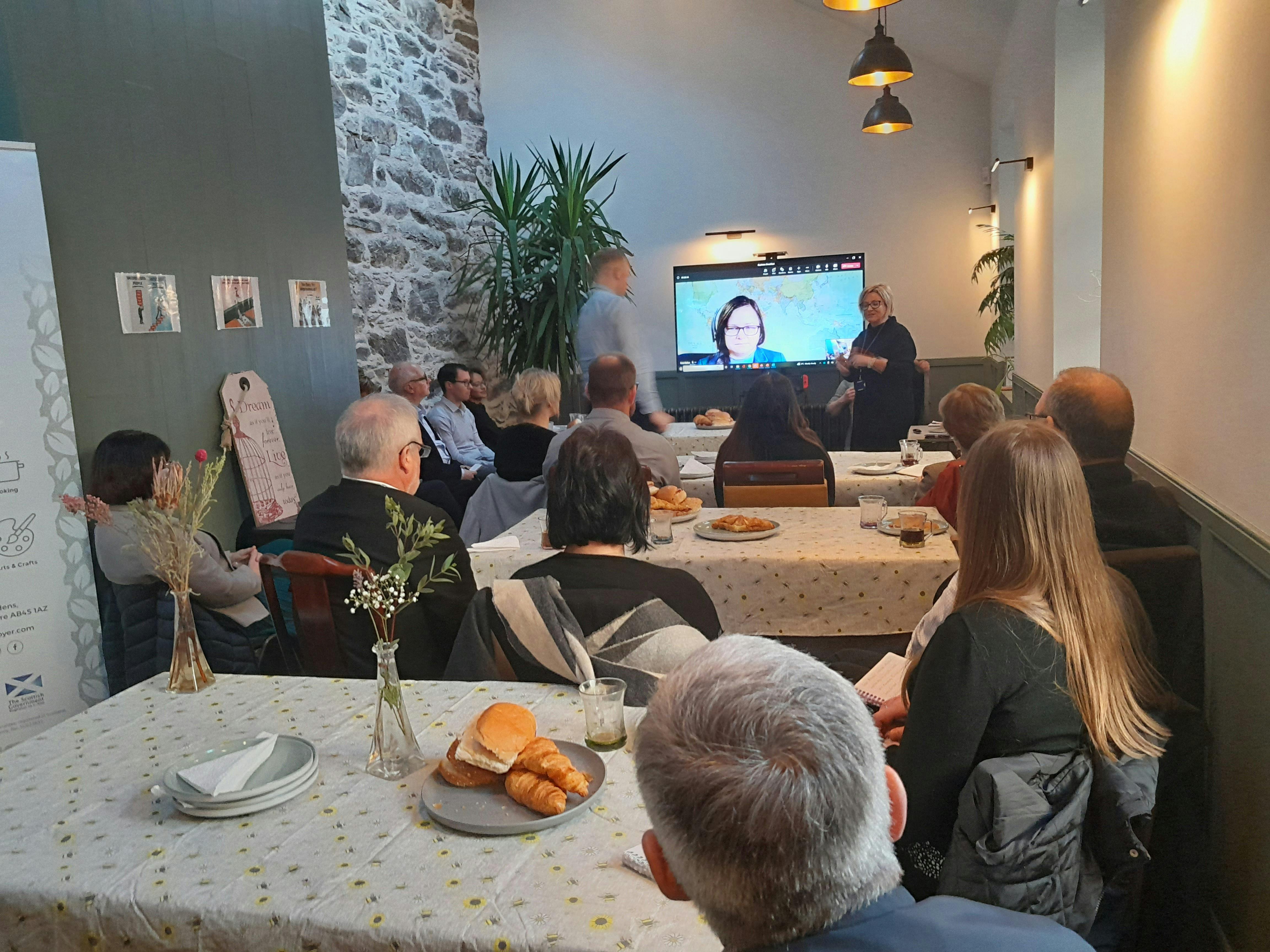 Picture shows attendees sitting at a table watching a presentation on screen