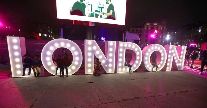 Art installation with neon letters that read London with people sitting in the letters and a screen above