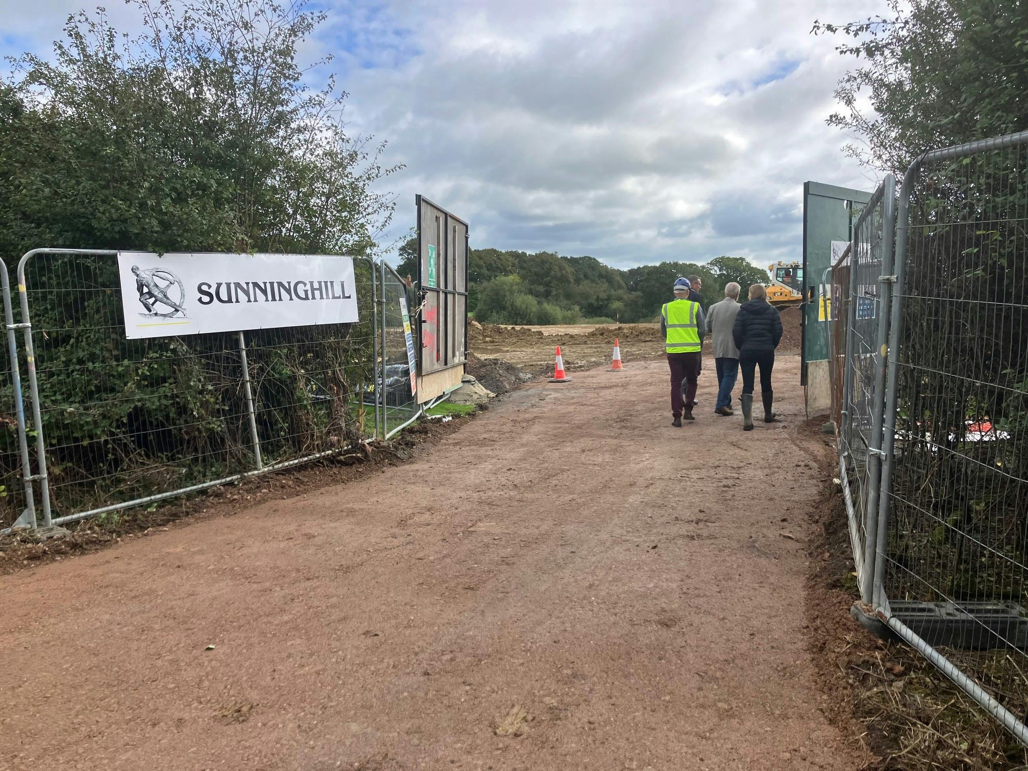 Shows a dirt track entrance to the building site that says Sunninghill on the fence