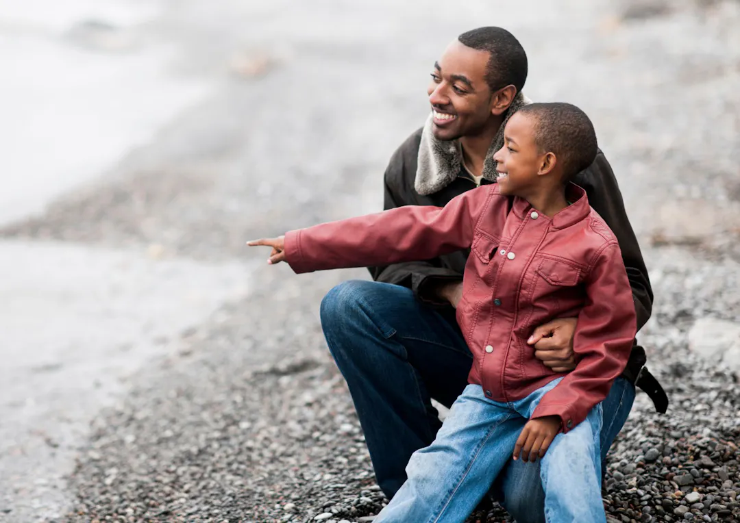 A smiling man and a young boy of Black British ethnicity sit together outdoors on a pebble beach. 
