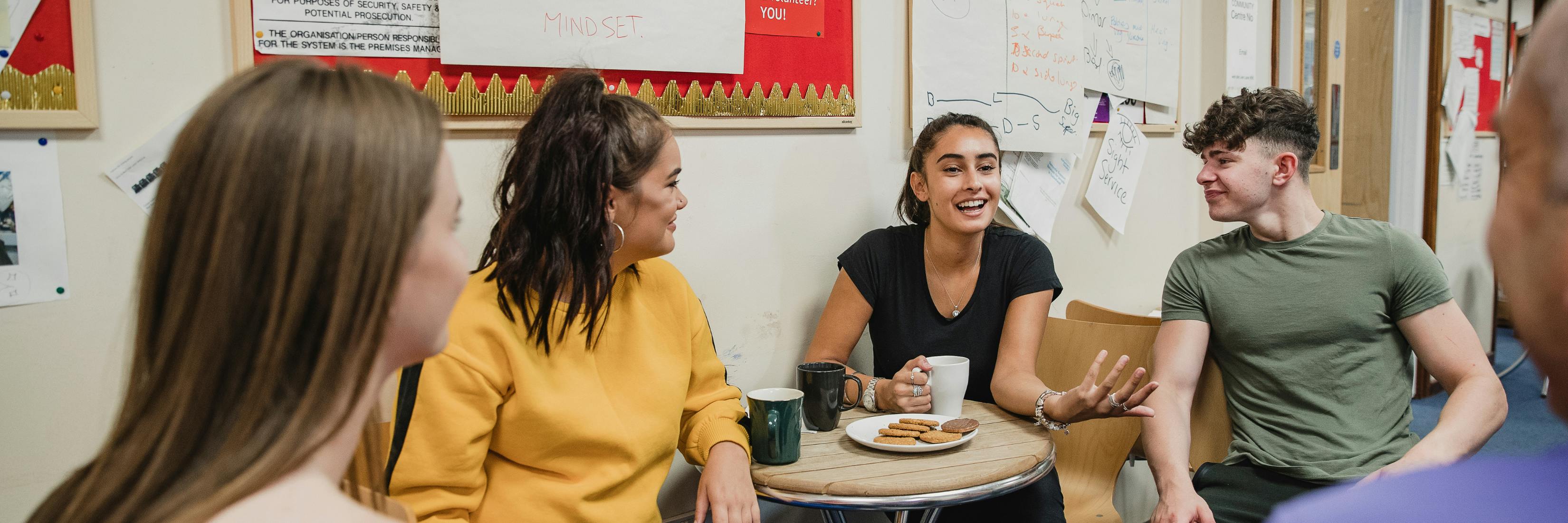 Image shows four young people talking around a table with hot drinks and biscuits