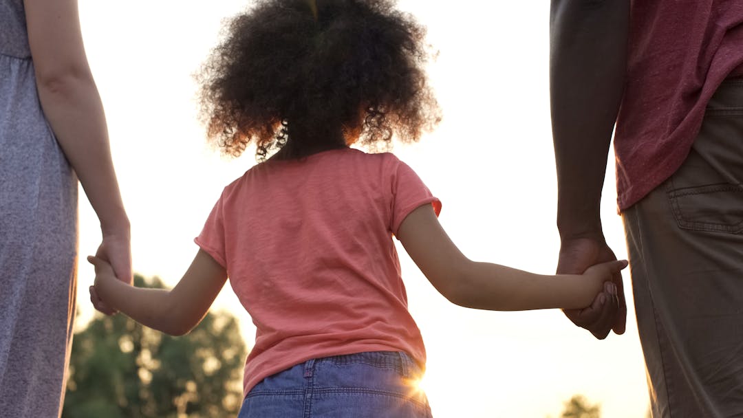 Child with curly hair looking towards sun, stood between two adults holding both their hands.