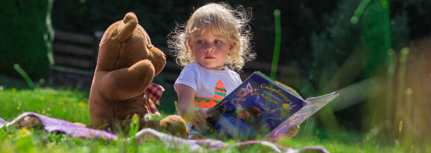 Young child sat on grass reading a book to a teddy