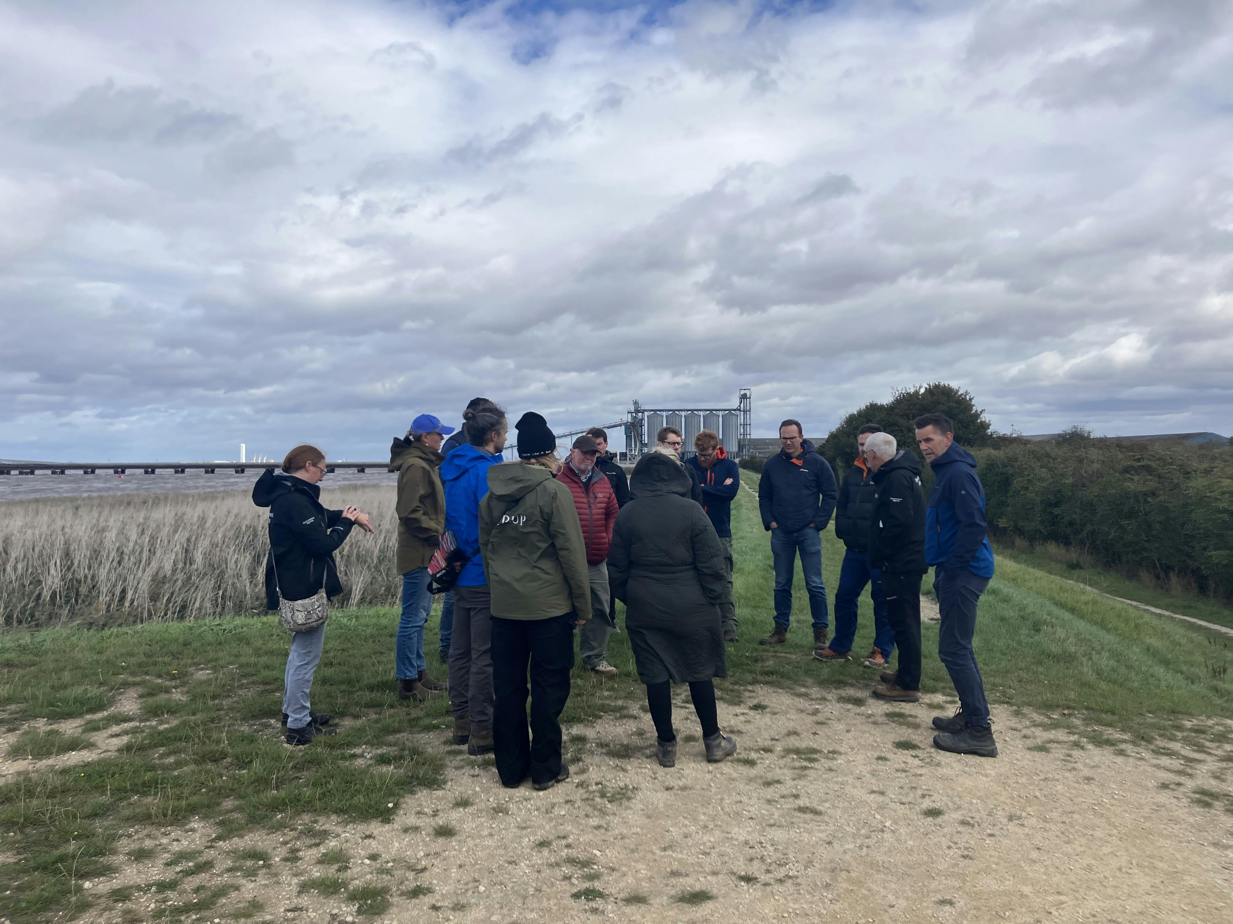 Photo of a group of people gathered on a grassy area with a dirt path under a cloudy sky, with an industrial or agricultural structure in the background. The group appears to be engaged in conversation.