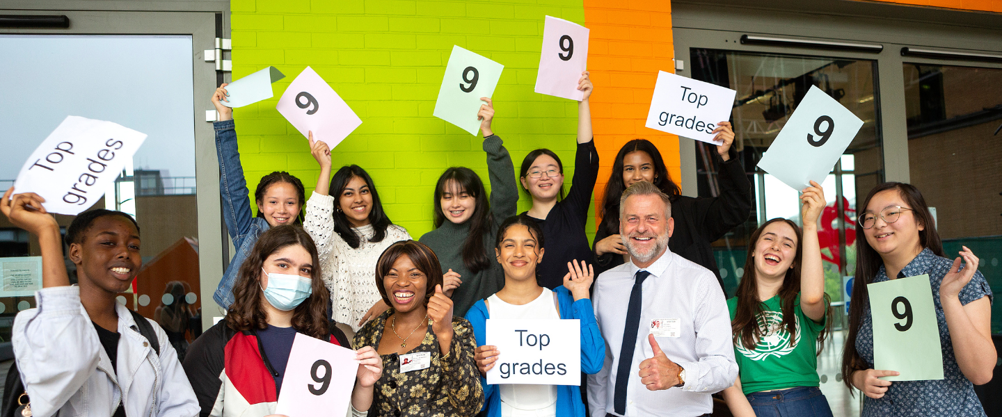 Group of smiling GCSE students on results day, holding signs that read 'top grades' and '9'