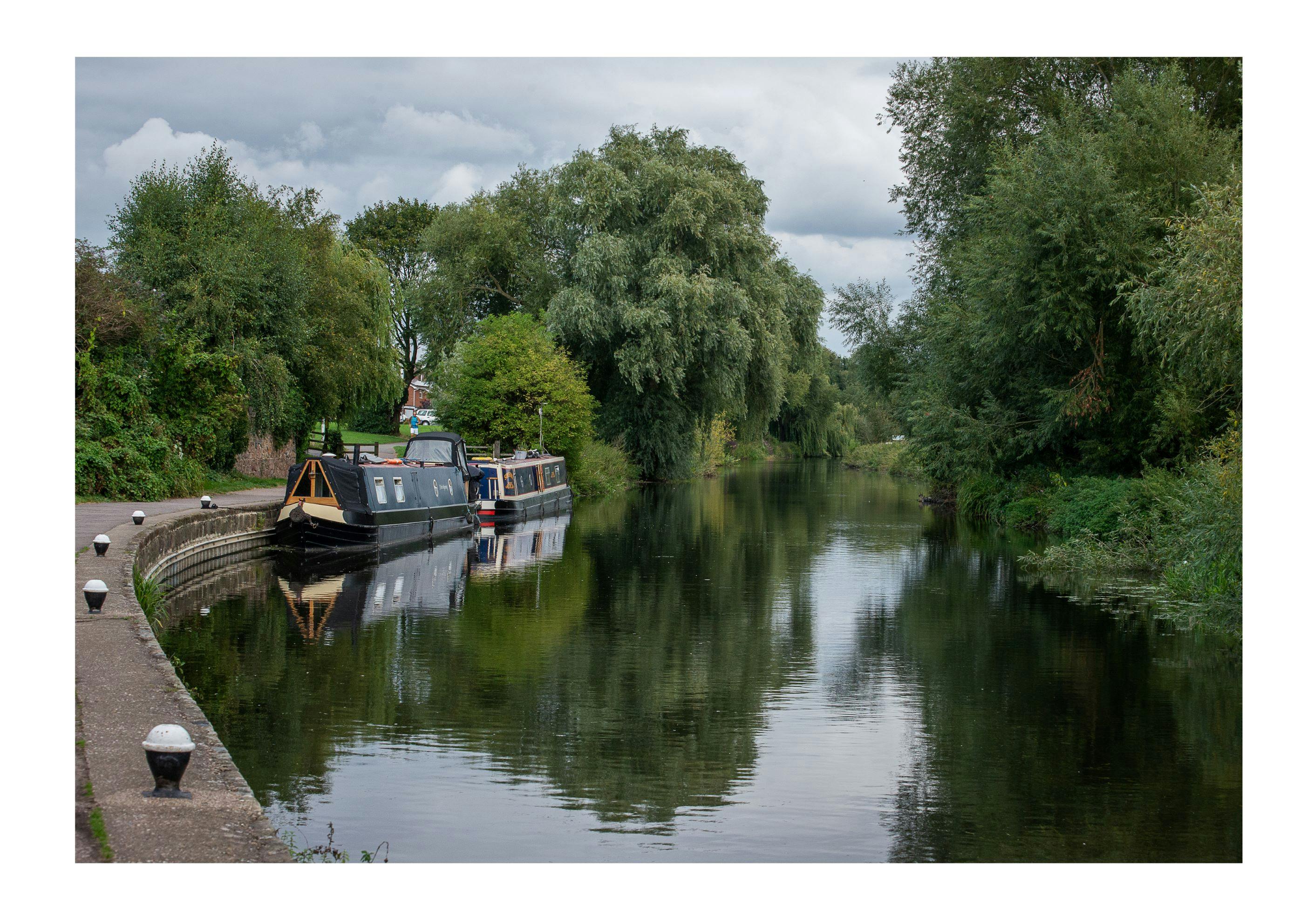 A photo looking along the River Soar near Aylestone Meadows. A couple of canal boats are moored alongside the tarmac footpath that runs along the left of the image.  Beyond the mooring area, the river is flanked continuously by trees.