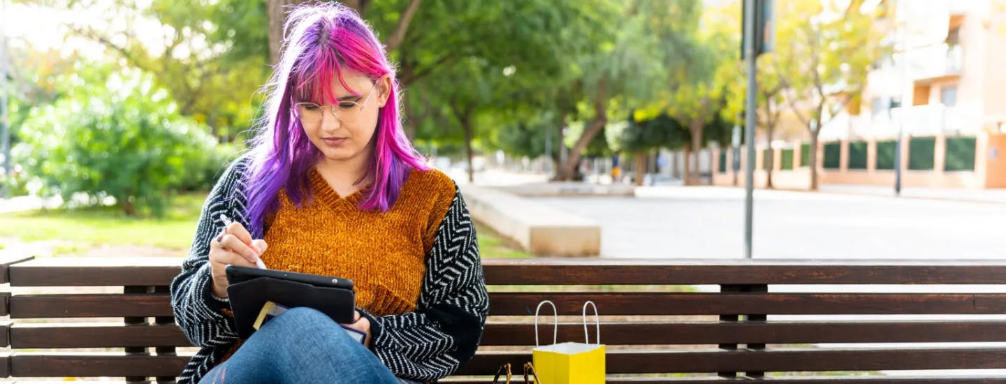 Young person with long purple hair and glasses, sitting outdoors on a bench, using a tablet.