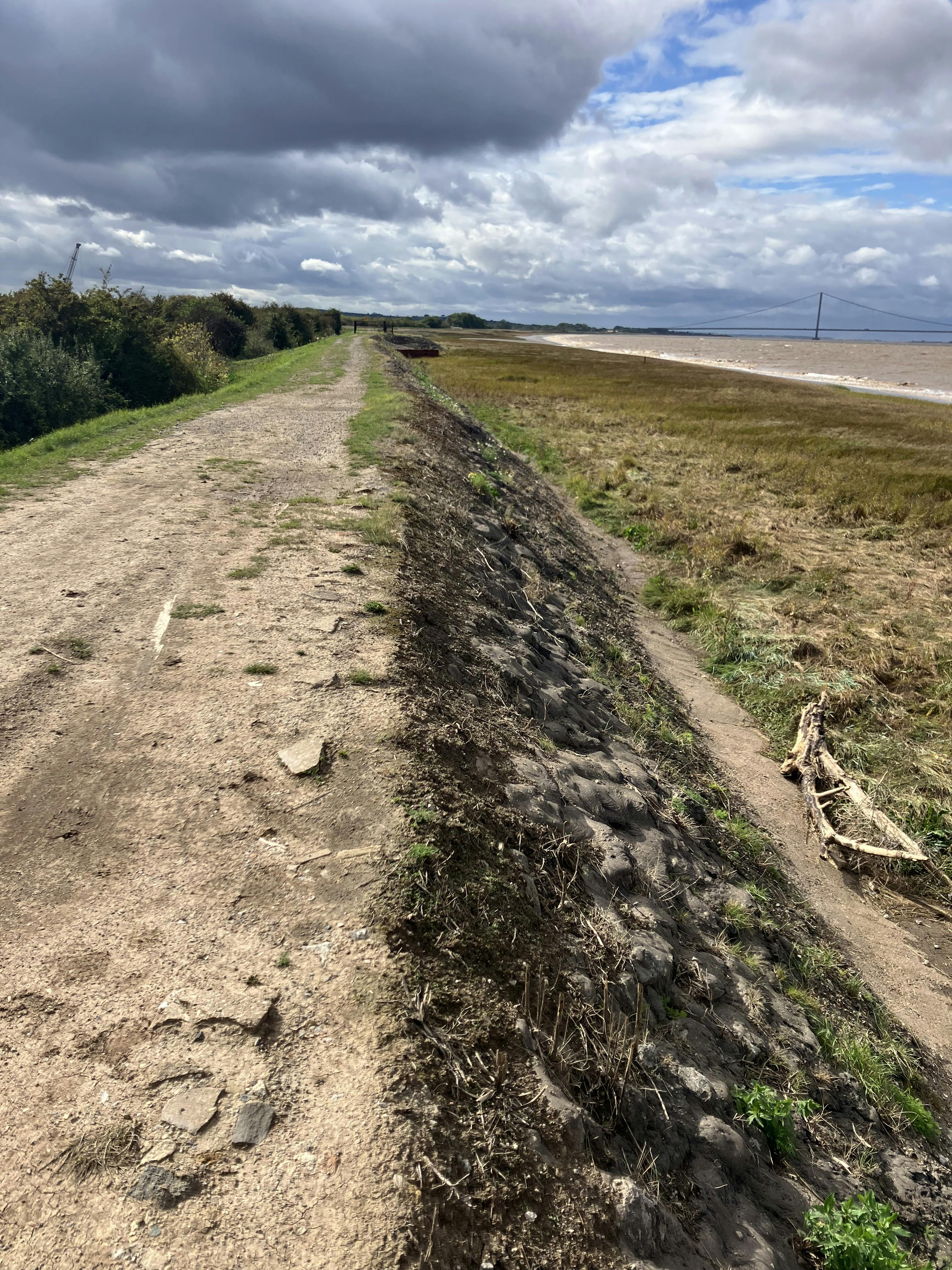 Photo of a dirt path running alongside a coastal marshland, bordered by vegetation and slightly elevated above the wetland. The sky is partly cloudy, creating a mix of light and shadow across the natural landscape.