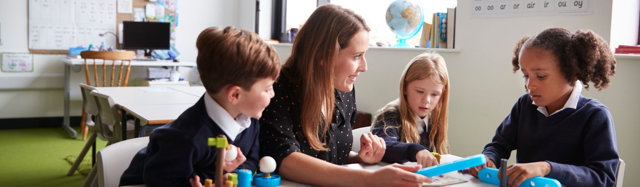 Primary school teacher sitting with pupils at table in a classroom.