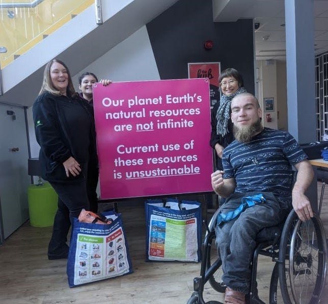 A group of people next to a pink sign with the wording "our planet Earth's natural resources are not infinite. Current use of these resources is unsustainable."