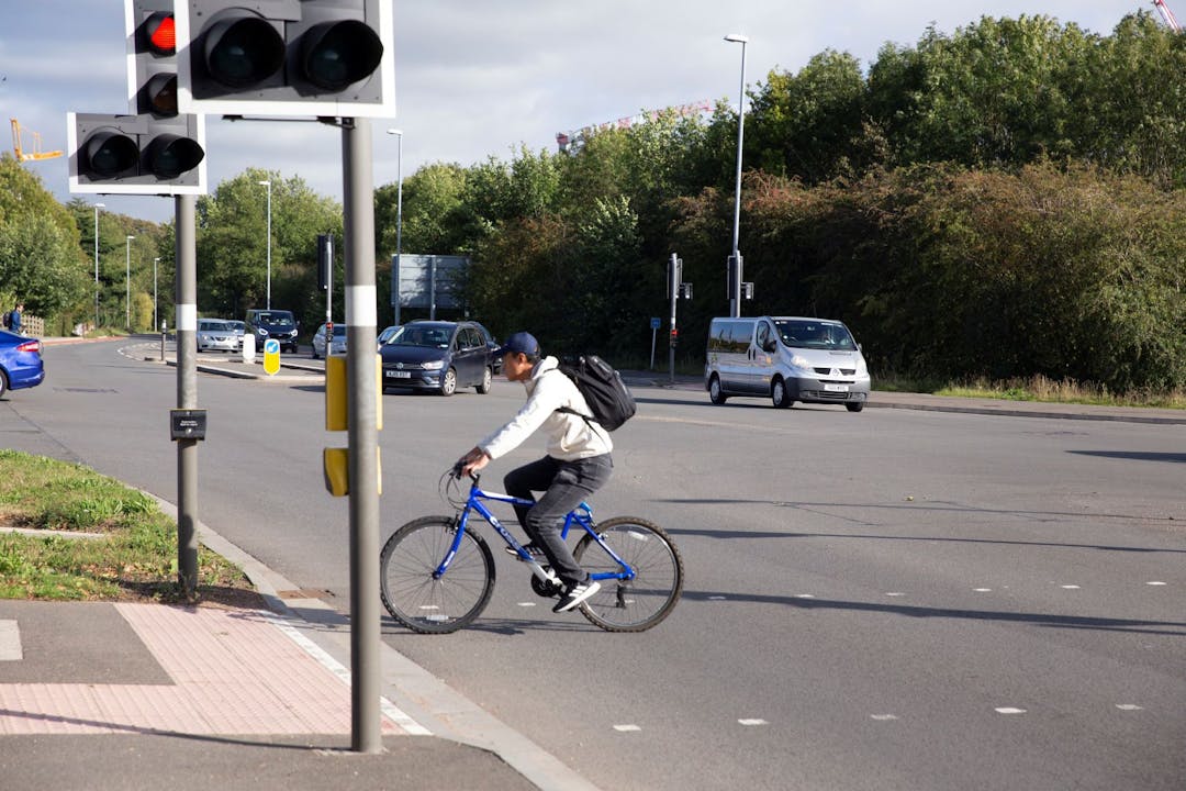 Man riding blue bicycle across crossing on Madingley Road with trees and traffic in the background