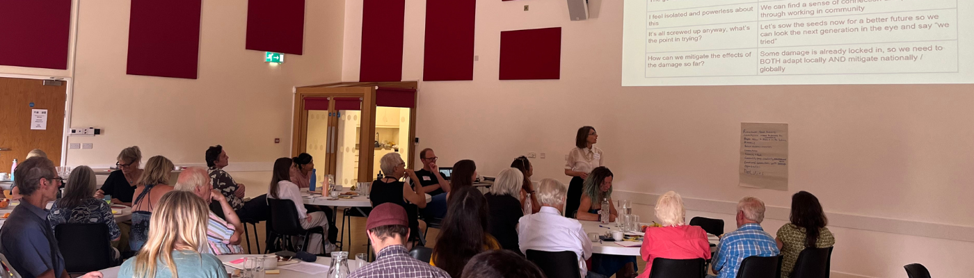 Image shows a presentation being delivered in a large hall with the audience sitting around tables listening