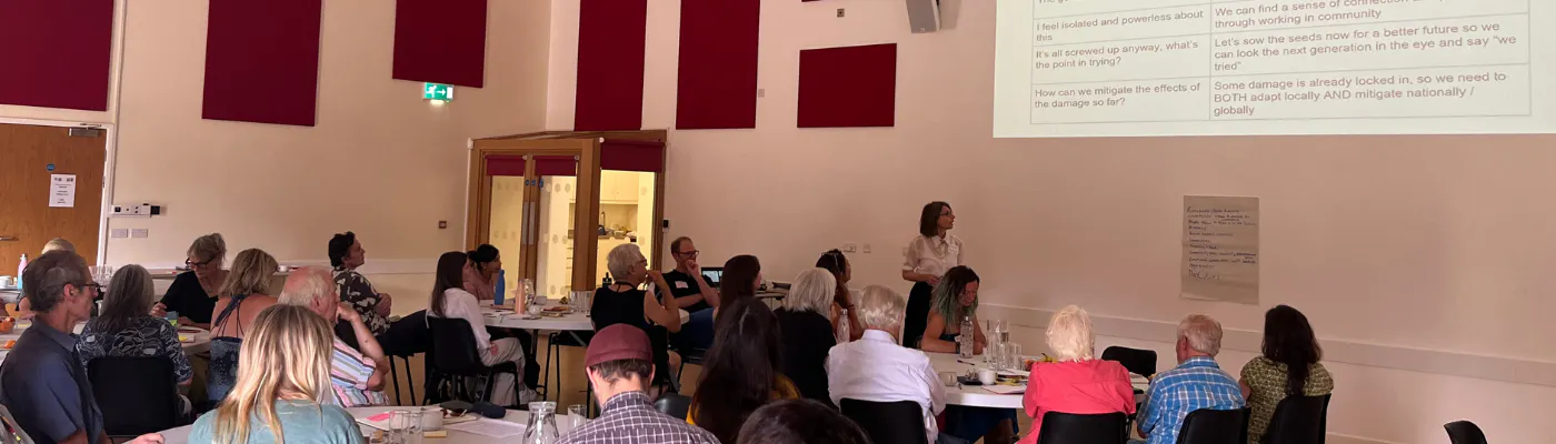 Image shows a presentation being delivered in a large hall with the audience sitting around tables listening