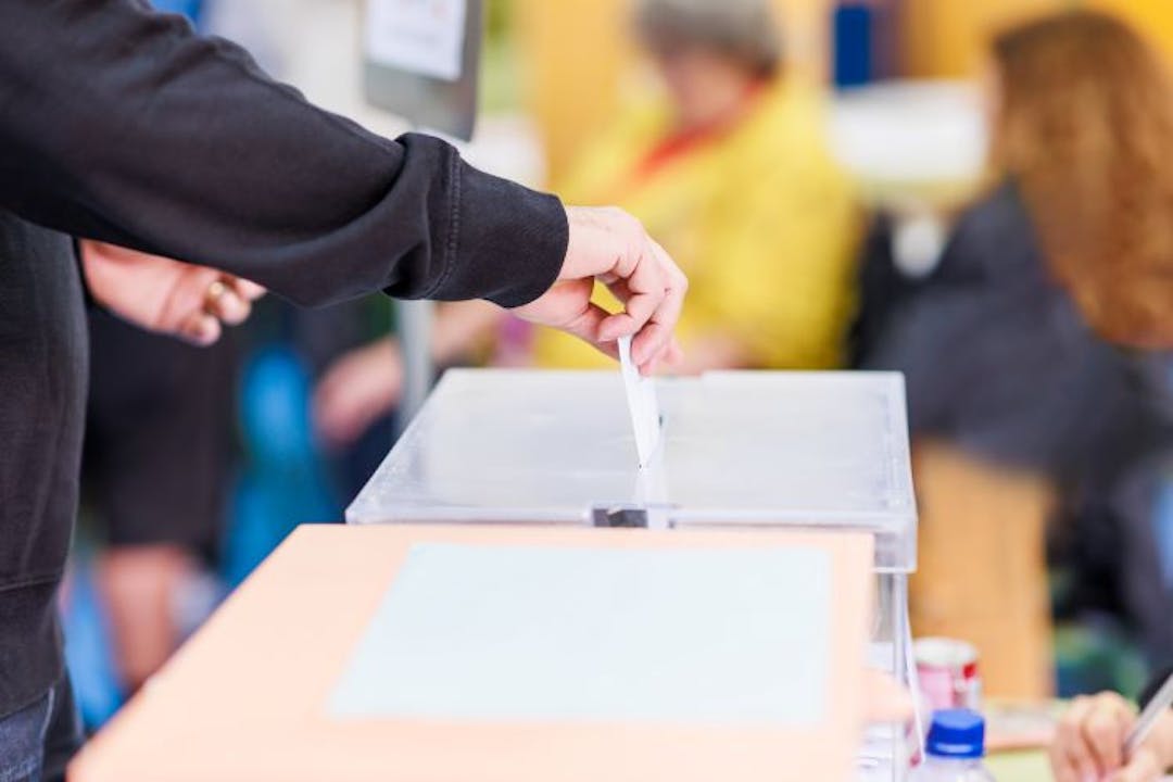 A persons hand putting a piece of paper into a voting ballot box banner