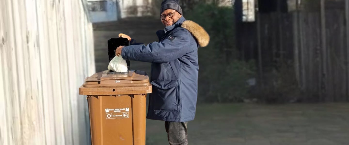 A man putting a small compostable bag of food waste into a food waste recycling wheelie bin outside a block of flats