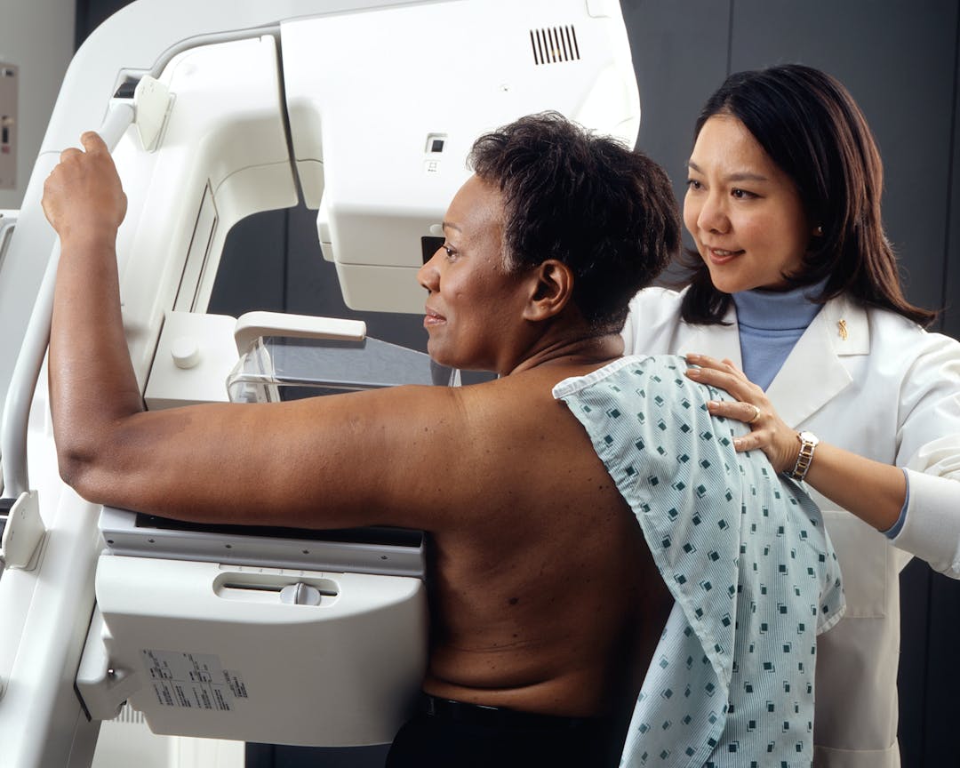 Woman Receives Mammogram.
 An Asian female technician positions an African-American woman at an imaging machine to receive a mammogram. Creator: Rhoda Baer