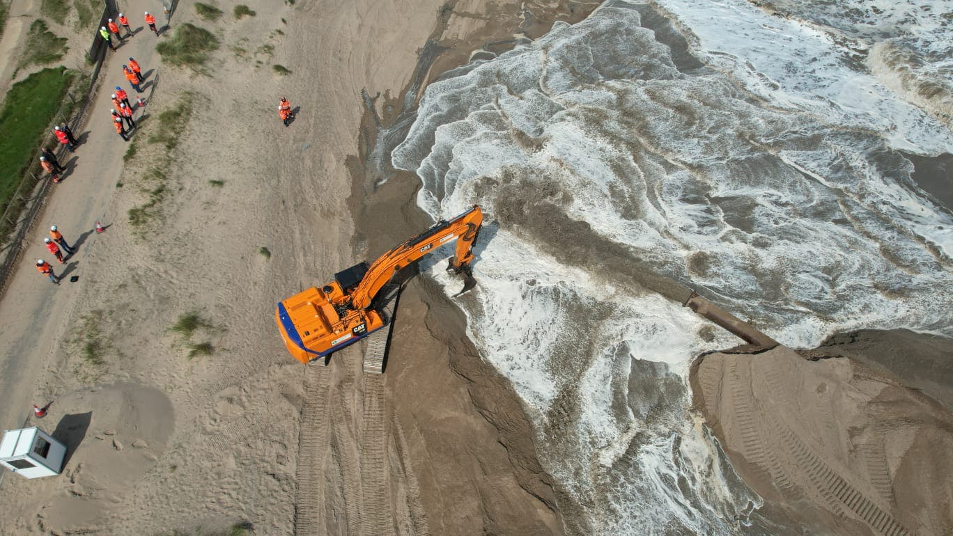 An excavator moves sand on the beach next whilst sand is pumped through a large pipe from the dredger