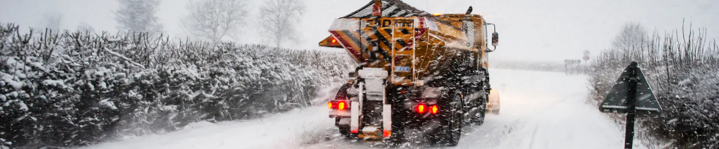 A snow plough driving along a snowy road whilst gritting