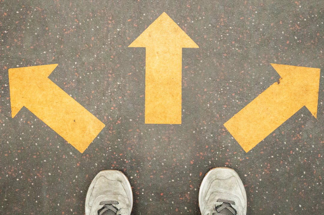 Overhead view of a pair of white trainers and 3 painted yellow arrows pointing at different directions against the road. 