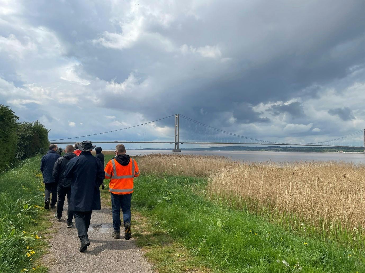 Photo of a group of people walking along a grassy path beside a body of water, with a large suspension bridge visible in the background under a cloudy sky.