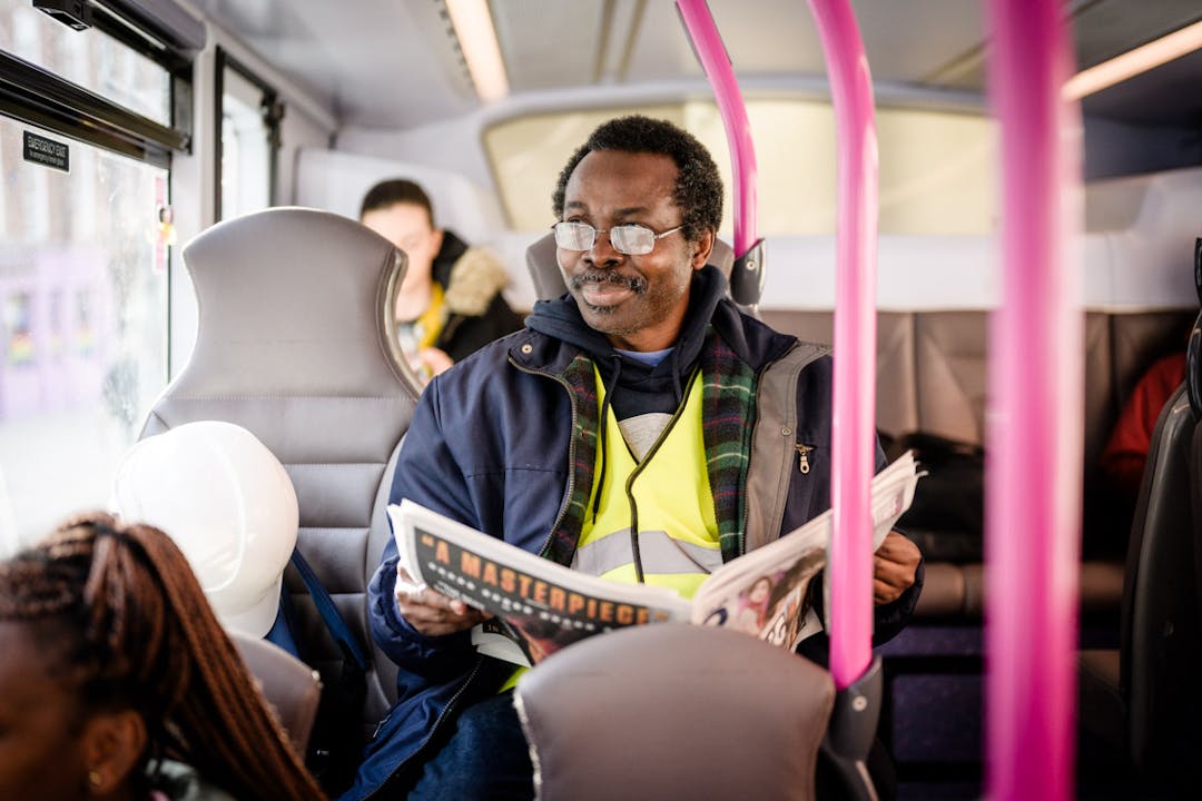 A man sitting on the bus. He has a newspaper open on his lap and is looking out of the window, looking contented. 