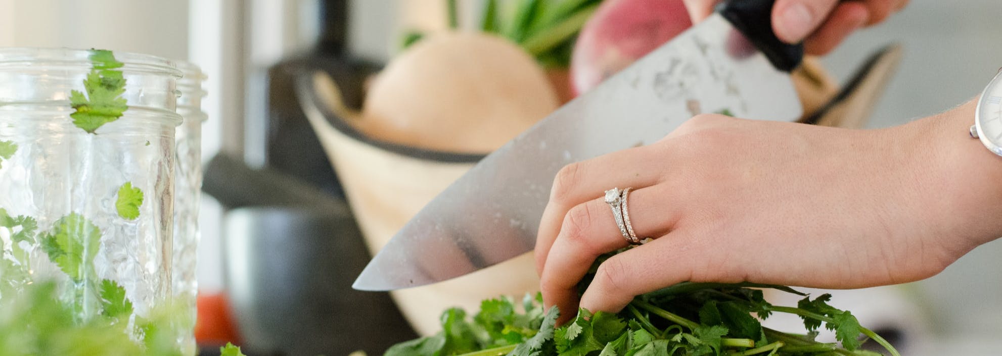 Close up of a pair of female hands chopping green herbs with a large knife with kitchen items in the background