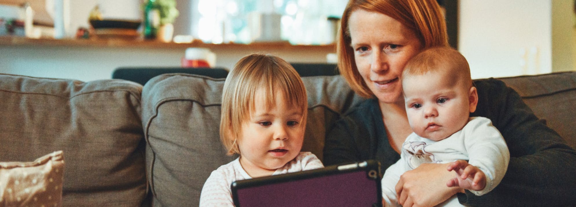 A woman, a toddler and a baby sitting on a sofa looking at a tablet