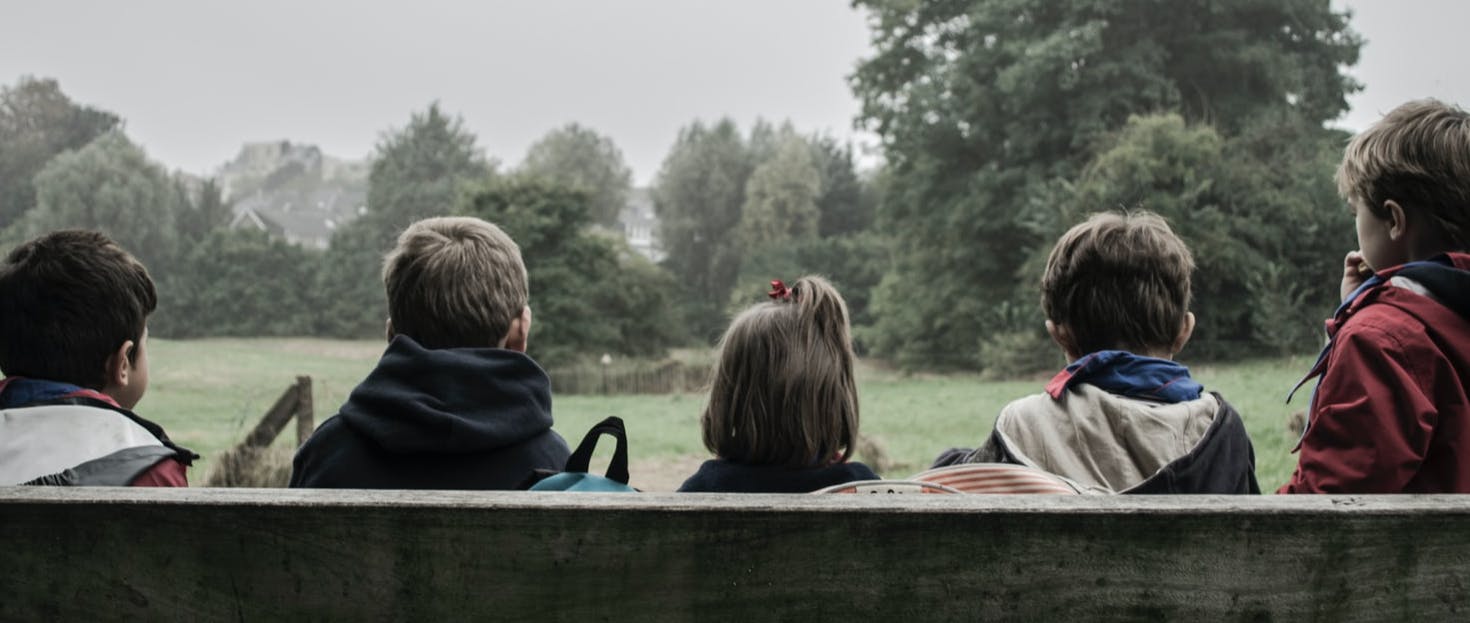 5 children sitting on a bench in the countryside. Facing away looking at trees in the background.