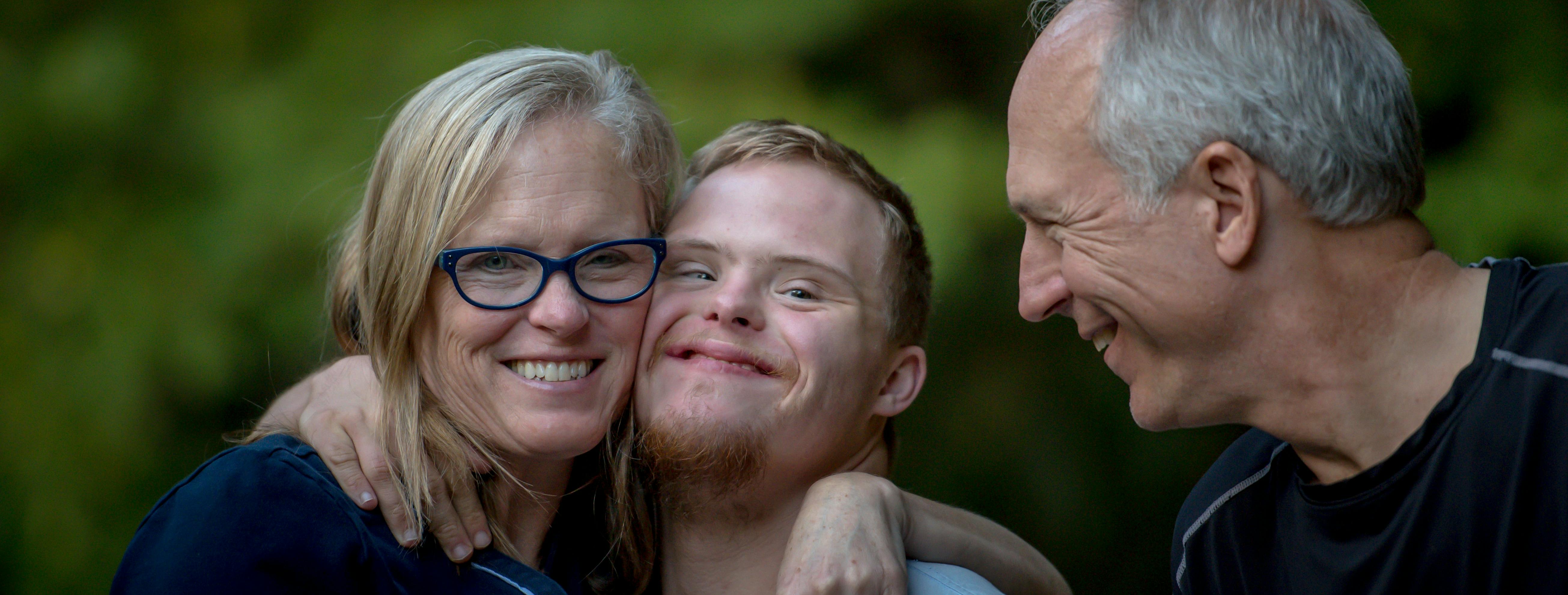 A young man with Down's Syndrome stands smiling with his parents