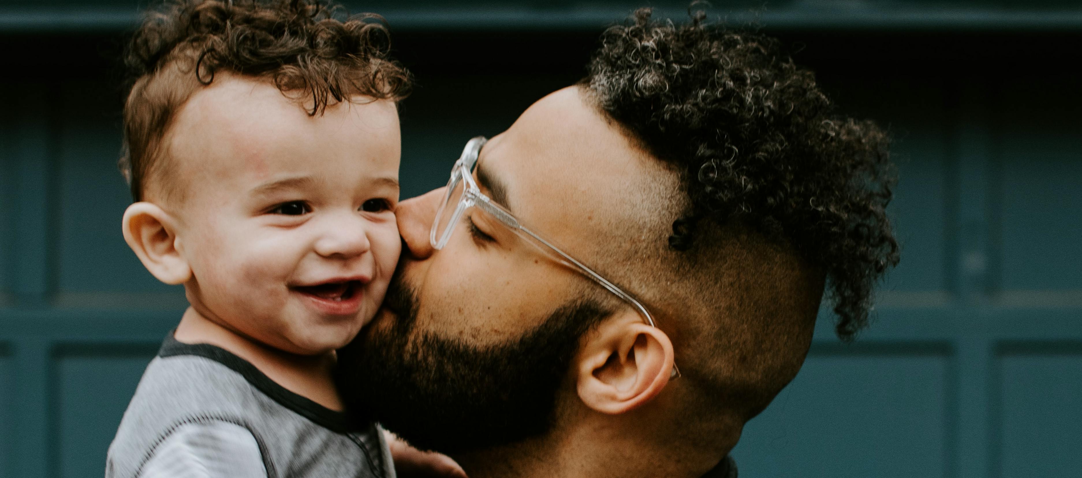 A little girl laughs as she covers the eyes of a smiling man in a black T-shirt. The man holds a gift.