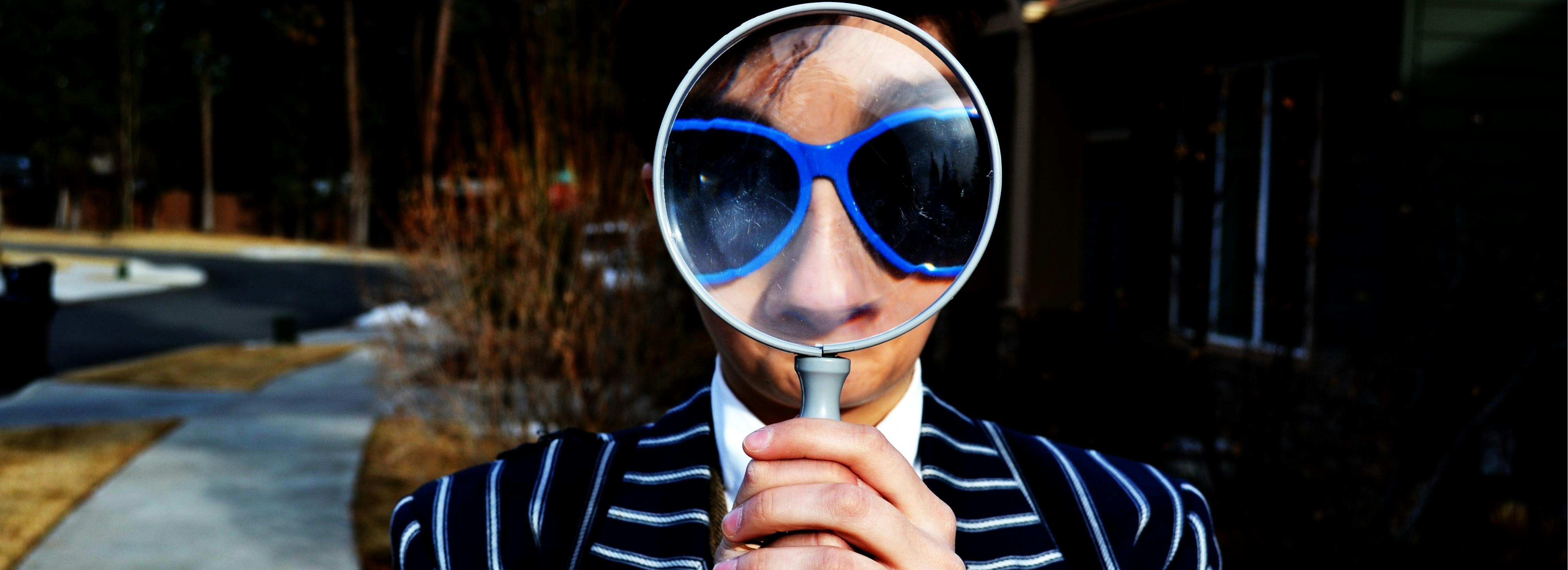 A person in a striped suit and sunglasses holds a magnifying glass in front of their face making their eyes very large