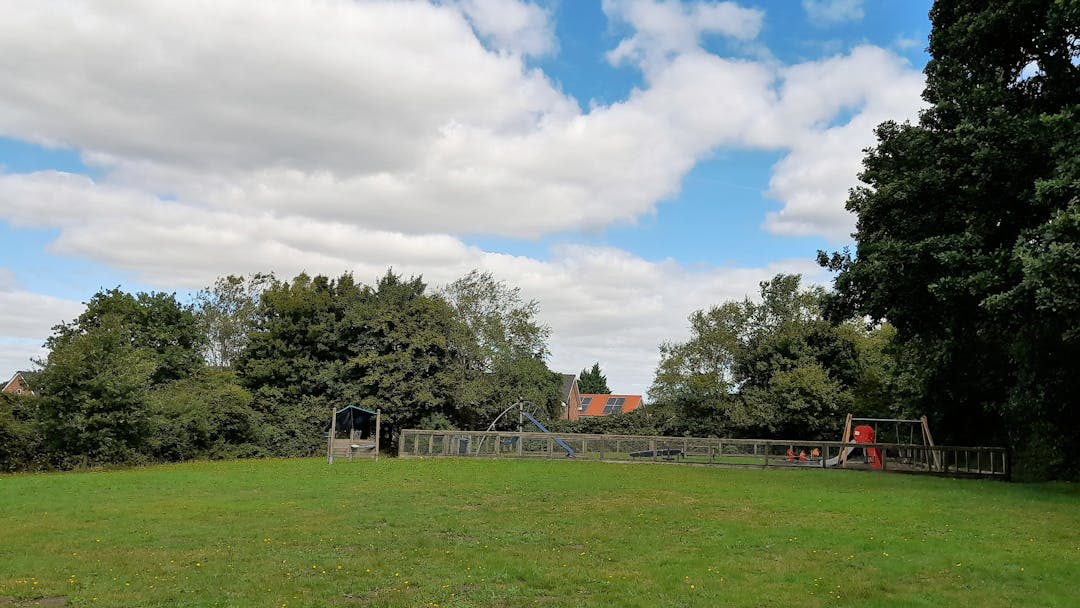 A play area with traditional play equipment, situated within a grass field bordered by mature trees