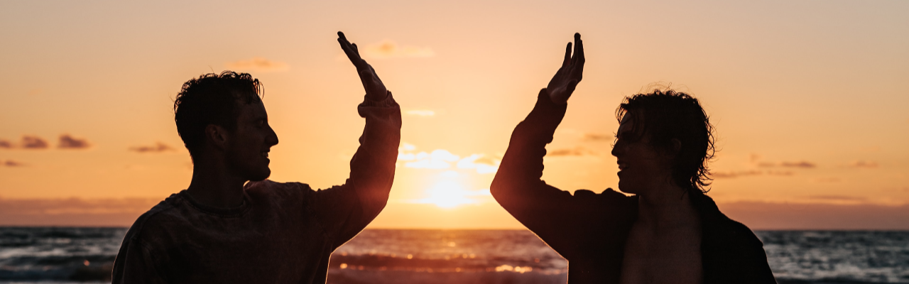 Silhouette of two men high fiving on a beach in front of a sunset