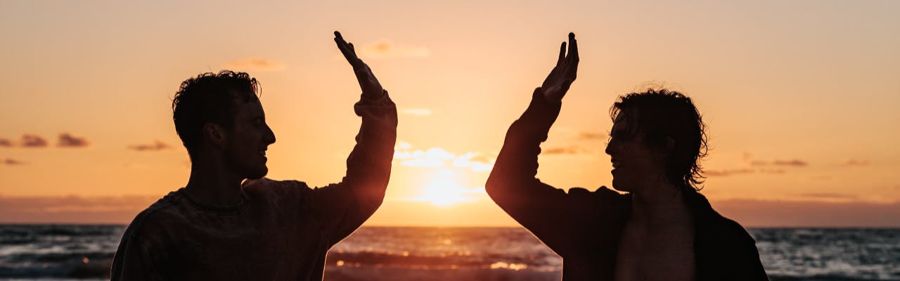 Silhouette of two men high fiving on a beach in front of a sunset