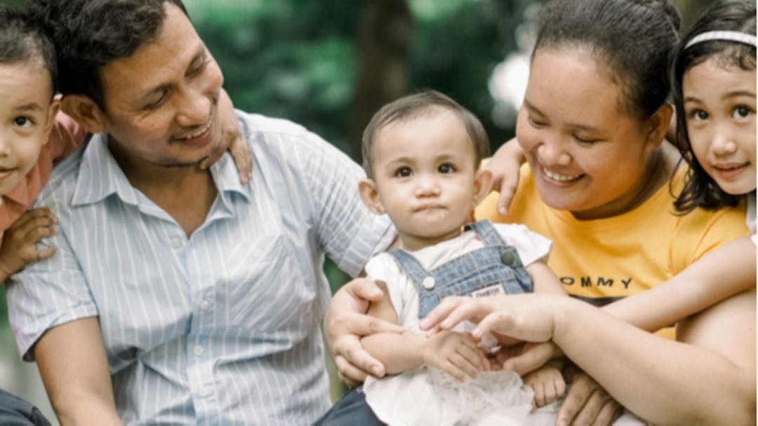 A family of Asian origin sitting outdoors. There is a mother, a father, and three young children.