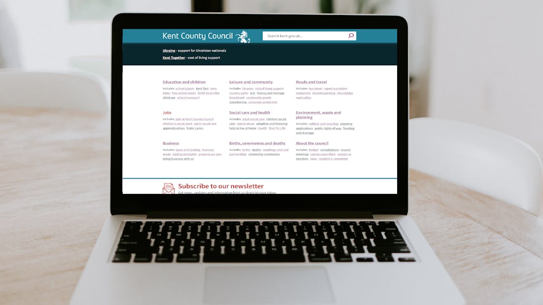 Image of a silver laptop sitting open on a wooden dining table, the screen shows a page on the kent.gov website 