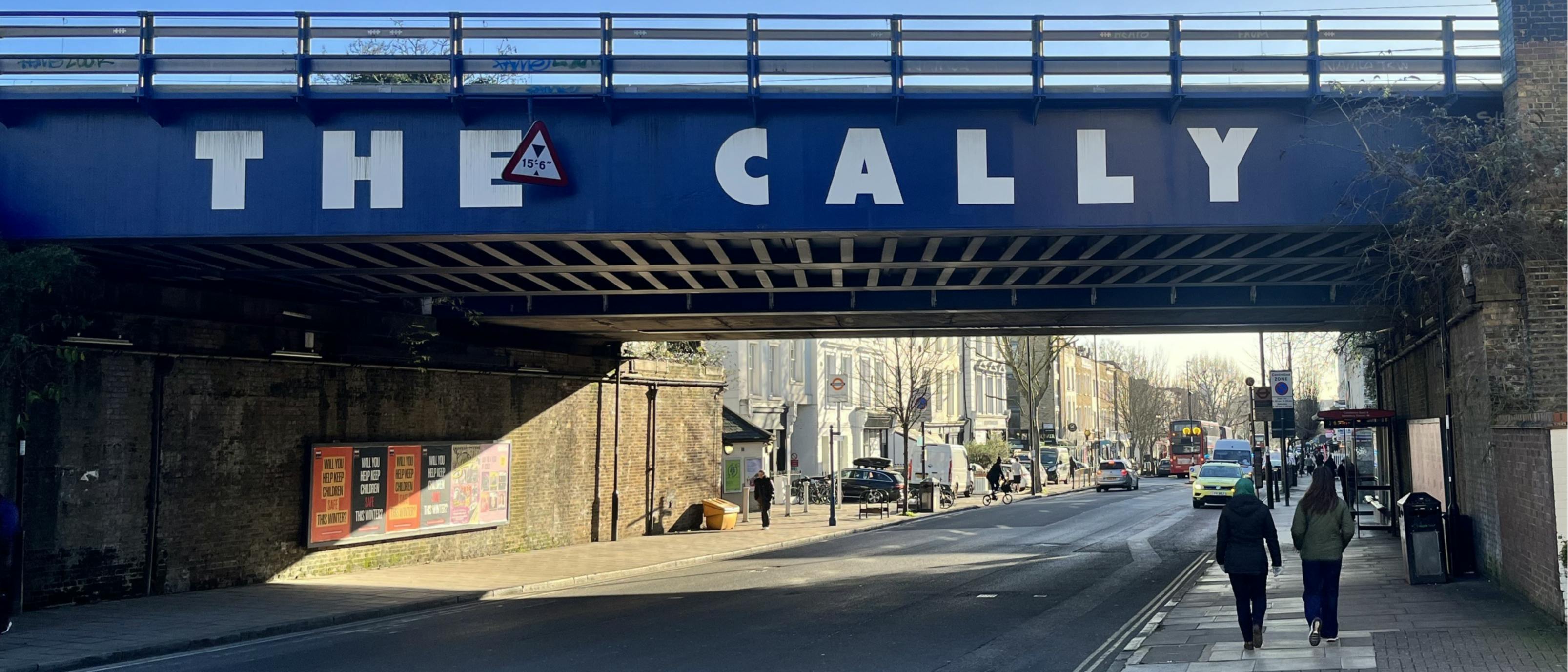 Caledonian Road at the junction with Freeling Street. Cars traveling in both directions. Tree over the hanging the road. 