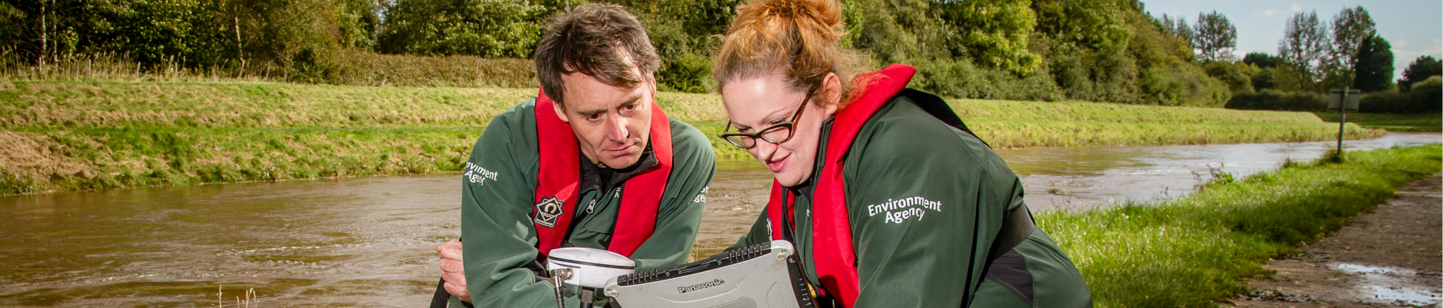 Two environment agency officers with testing equipment by a river