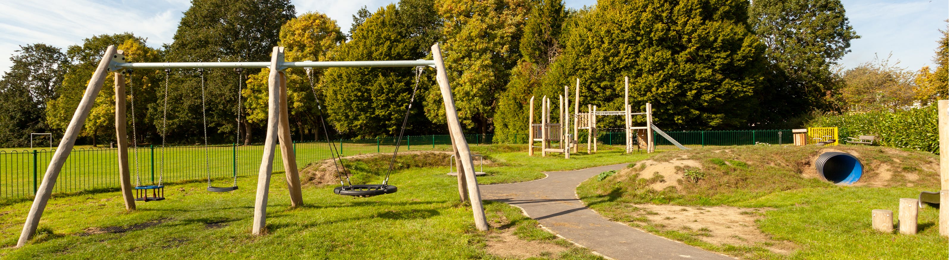 Play equipment including swings, a tunnel and climbing frame. All on green grass surrounded by mature trees.