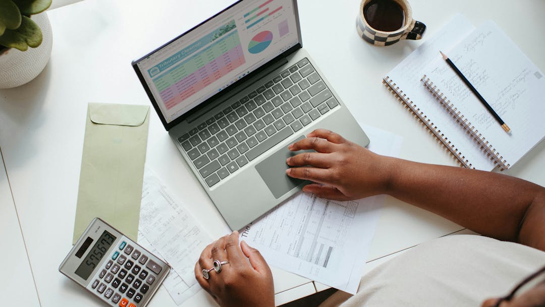 A birds eye view of a person working on a laptop which is surrounded by notebooks, a calculator and a cup of coffee