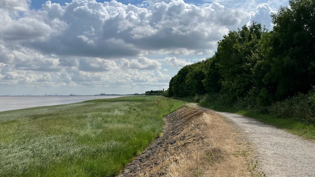 Photograph of scenic landscape featuring a gravel path beside a calm body of water, bordered by lush green grass on one side