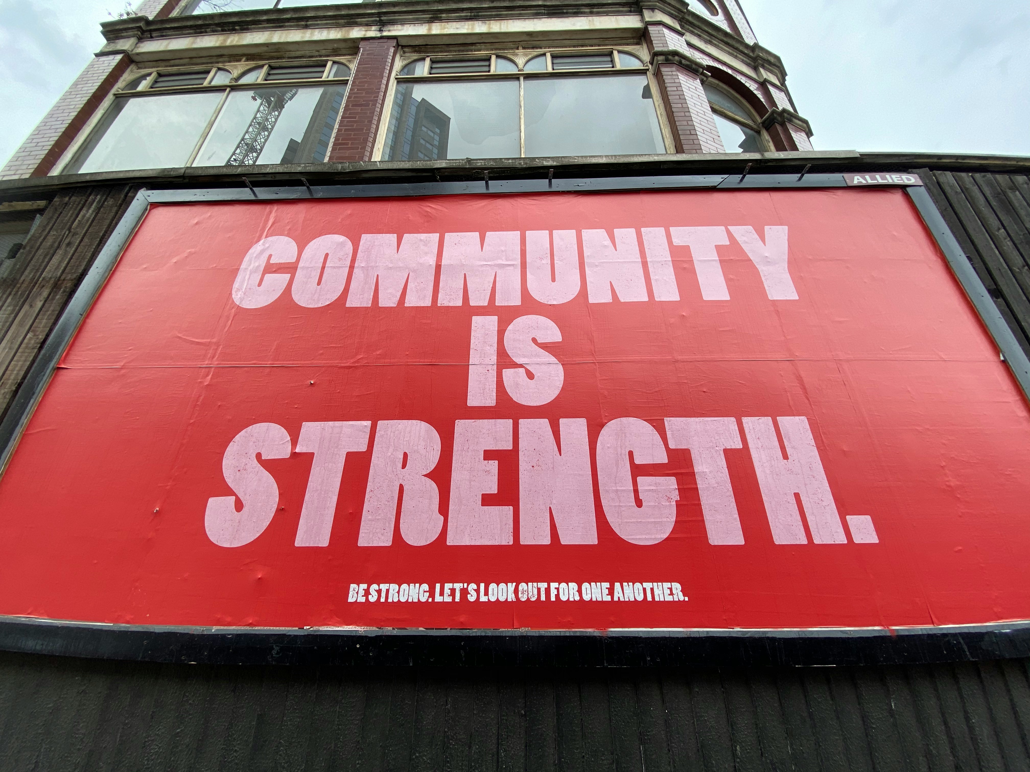 Red billboard with  Pink Letters reading Community is strength in smaller letters be strong lets look out for one another