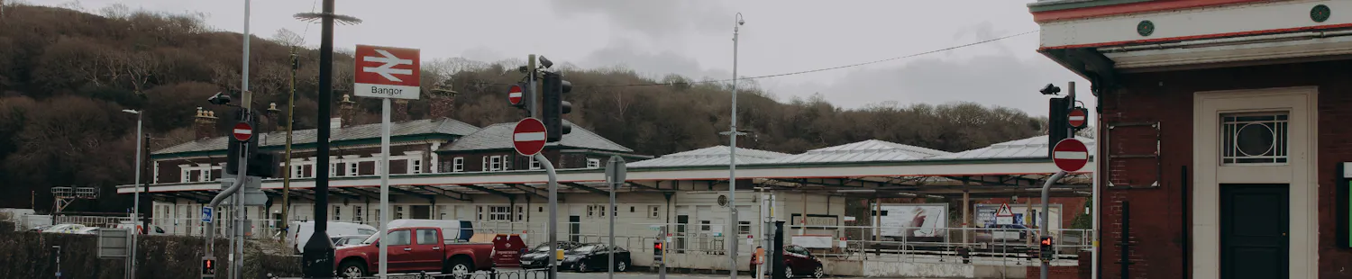 Cwrt blaen gogleddol Gorsaf Bangor, yn dangos cyffordd priffordd darmac a maes parcio gydag arwyddion Gorsaf Bangor.