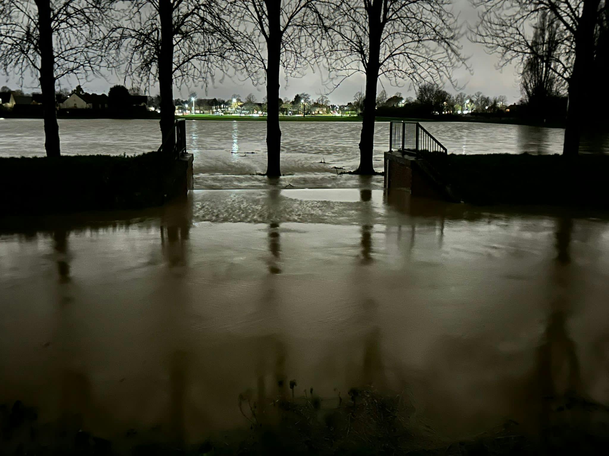 A dark photo taken at night of water flowing across a spillway, which is a gap between two sections of embankment, from the brook out into the playing field area that forms one of the flood storage cells.