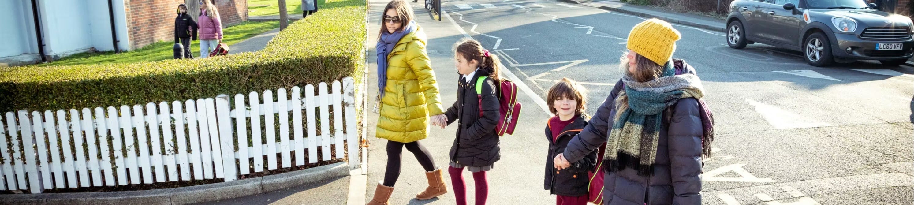 A group of children and an adult walking together on a residential street, crossing near a white picket fence.