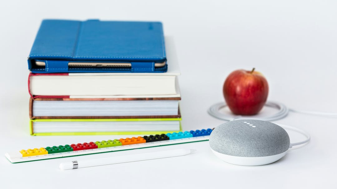 Books and an ipad in a stack, an apple, a pencil and a ruler.