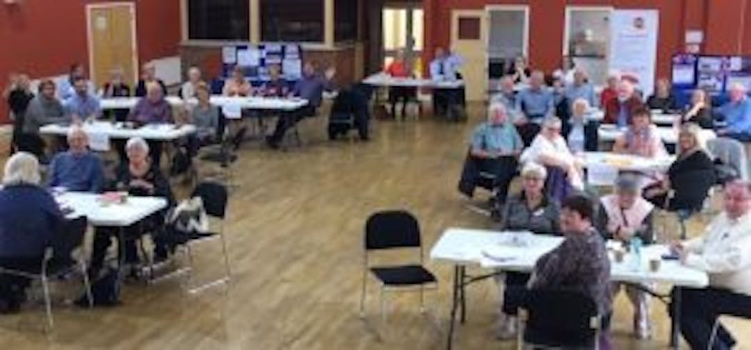 Patient Participation Group (PPG) Network meeting in action at Churchdown Community Centre. Six tables of PPG members discussing how they work together. There are, on average, six PPG members on each table and it is a mix of males and females.