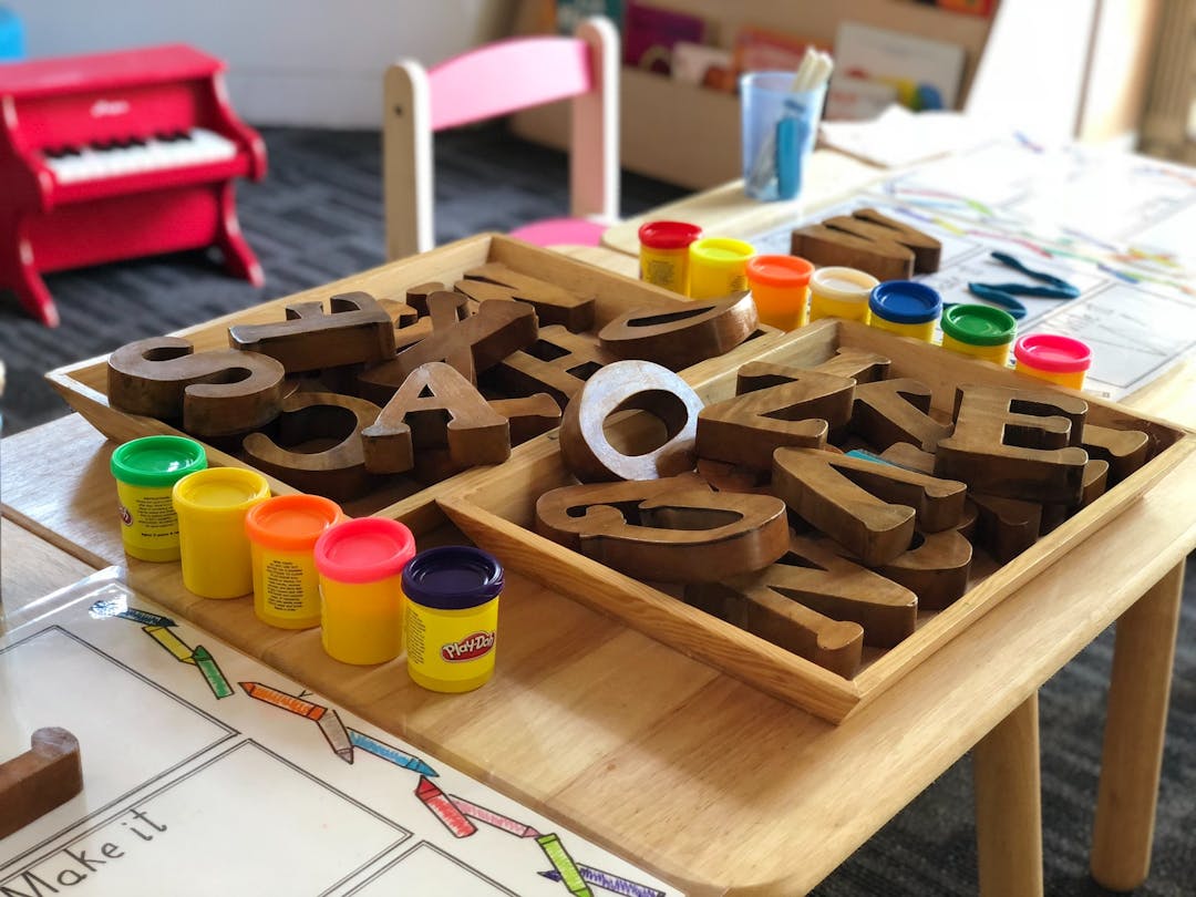 A table has play doh and wooden letters on it, childs toys can be seen in the background