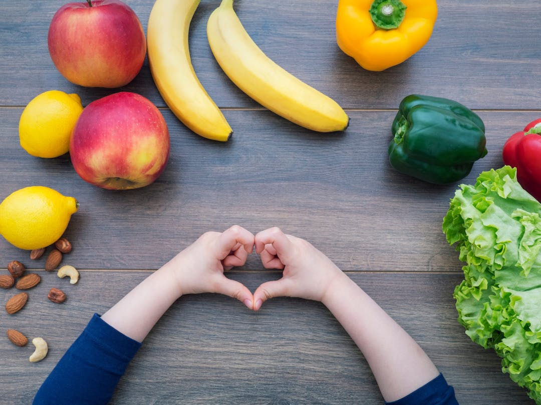 Image of a range  of fruit and vegetables (apples, lemon, banana, peppers, lettuce and nuts) laid out on a table with a child making a heart shape with their hands indicating her love of fresh fruit and vegetables