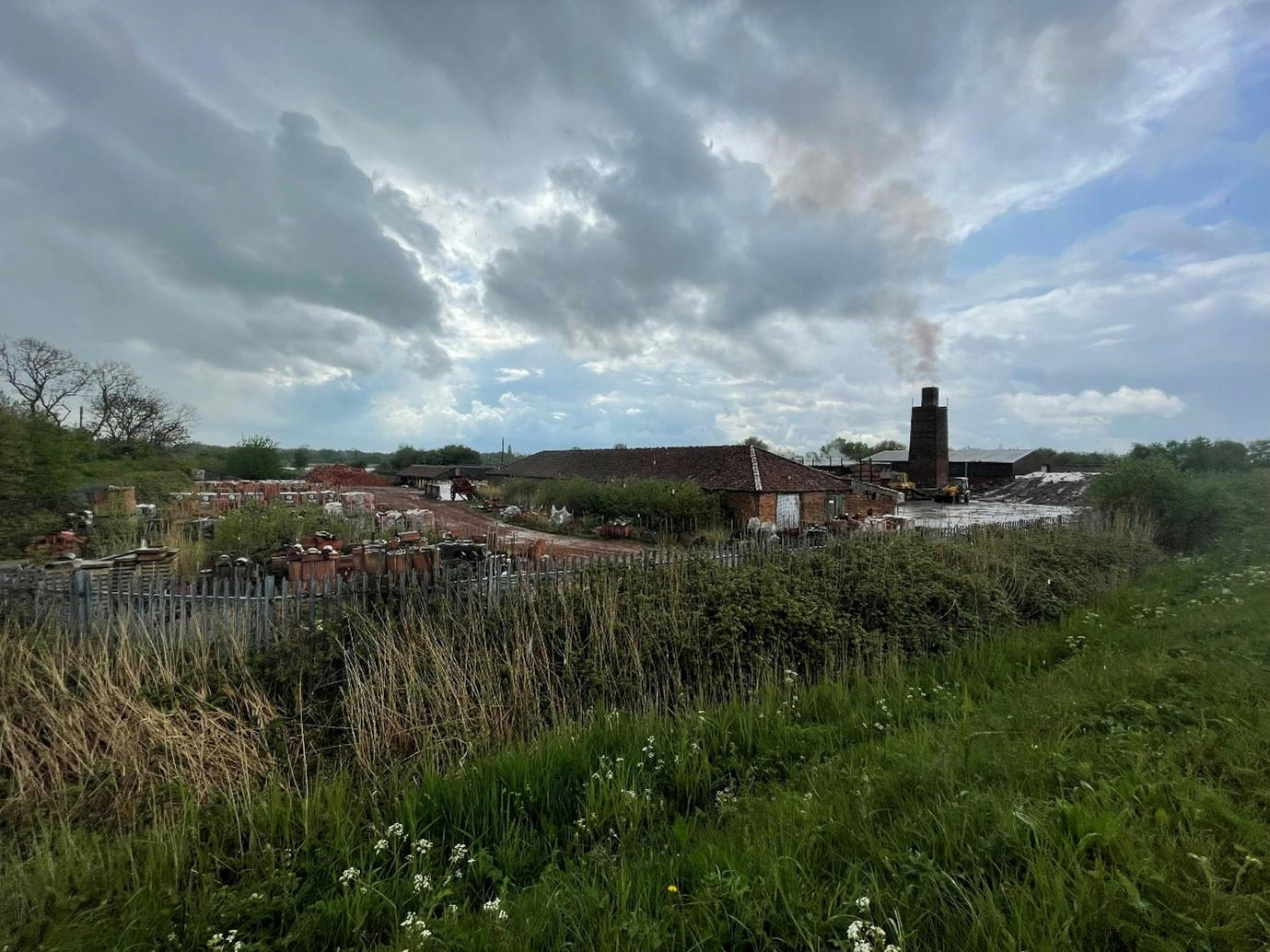 Photo of a tall brick chimney emitting smoke with wild grasses and flowers in the foreground. Beyond the fence, stacks of bricks and construction materials are visible.